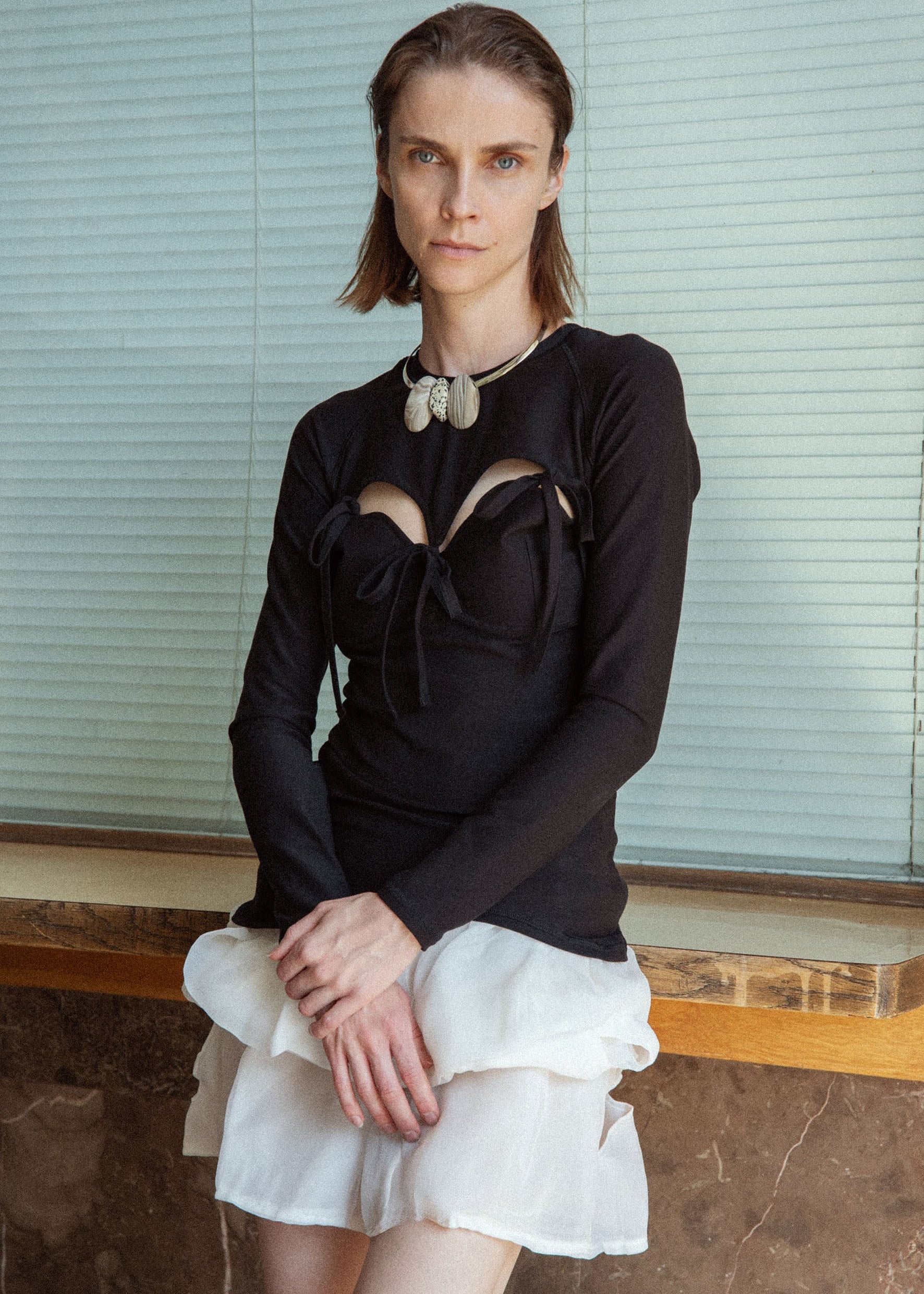 Woman wearing a black long-sleeve top with cut-out design and a white skirt against a neutral background.