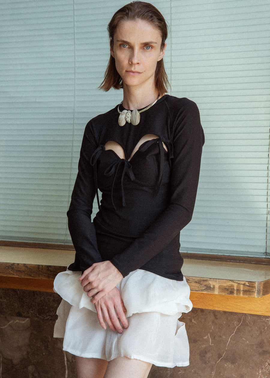 Woman wearing a black long-sleeve top with cut-out design and a white skirt against a neutral background.