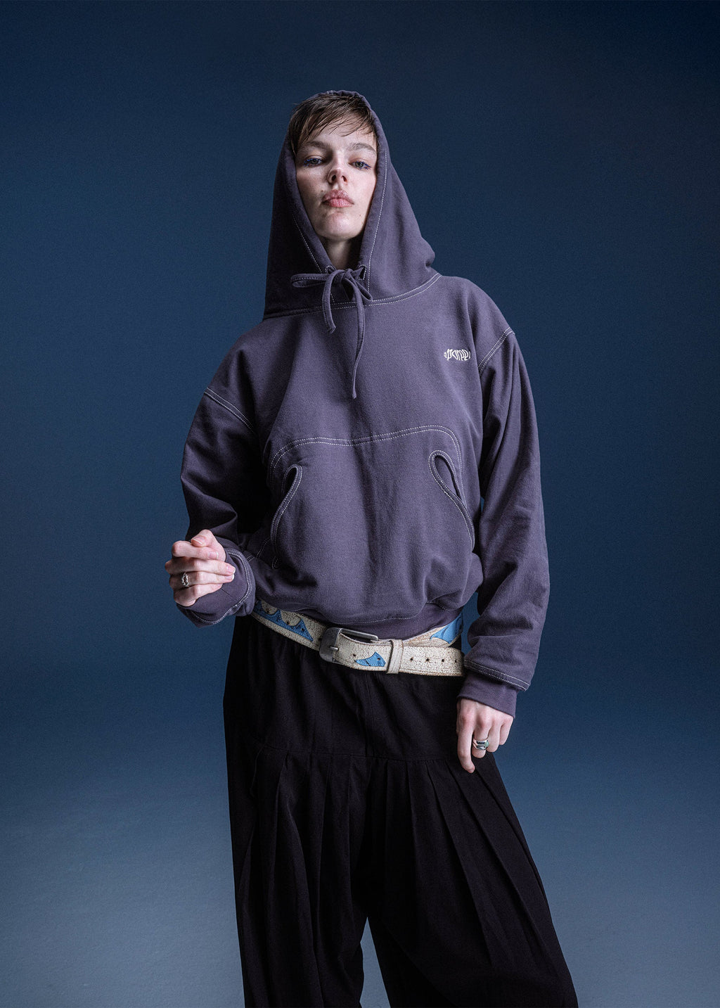 Person wearing a dark hoodie and black pants against a blue background