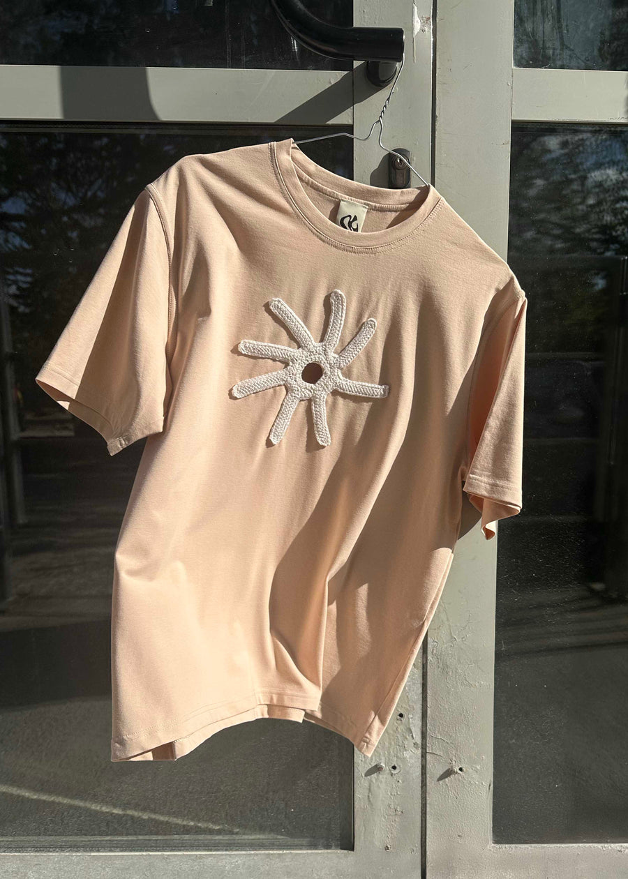 A nude-colored t-shirt with a decorative crochet applique in the form of a star on the chest, displayed on a hanger against a glass door.